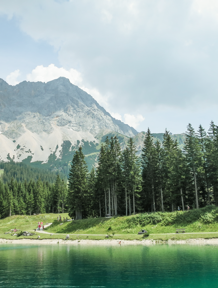Bergsee mit spiegelnder Wasseroberfläche vor bewaldeten Hängen und Gipfeln.