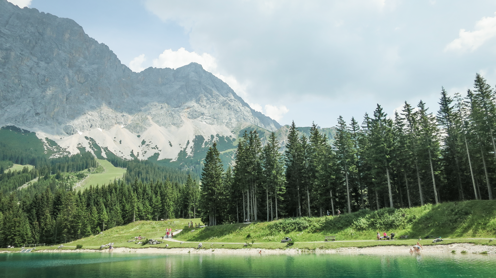 Bergsee mit spiegelnder Wasseroberfläche vor bewaldeten Hängen und Gipfeln.