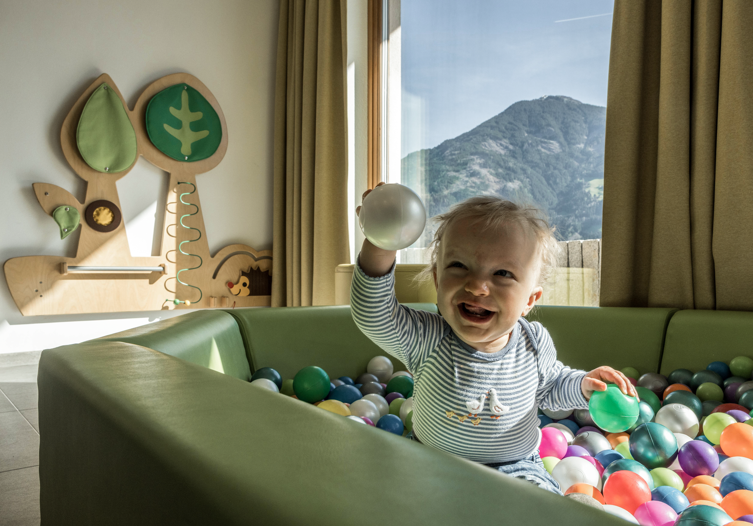 Kleinkind spielt in einem Bällebad vor einem großen Fenster mit Bergblick.