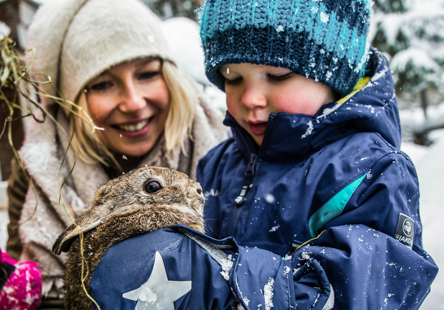 Erwachsene Person und Kind halten gemeinsam ein Kaninchen im Arm in einer winterlichen Schneelandschaft.