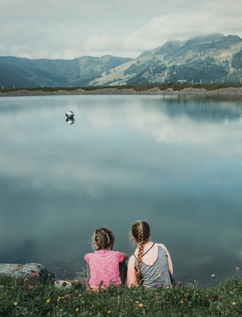 Zwei Personen sitzen auf einer Wiese und genießen den Blick auf einen Bergsee.
