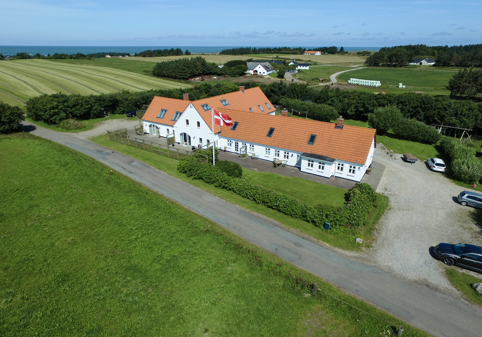 Schräg aufgenommene Ansicht eines langen Bauernhauses mit rotem Dach, neben einer Straße und Wiesen