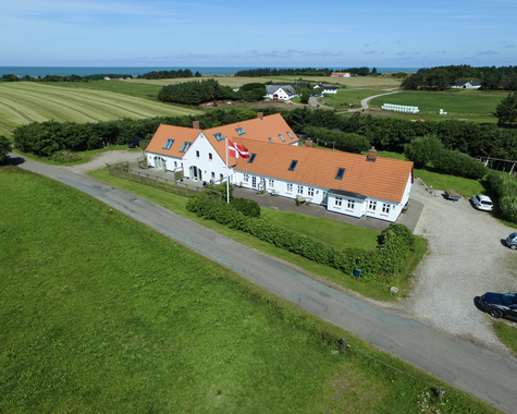 Schräg aufgenommene Ansicht eines langen Bauernhauses mit rotem Dach, neben einer Straße und Wiesen