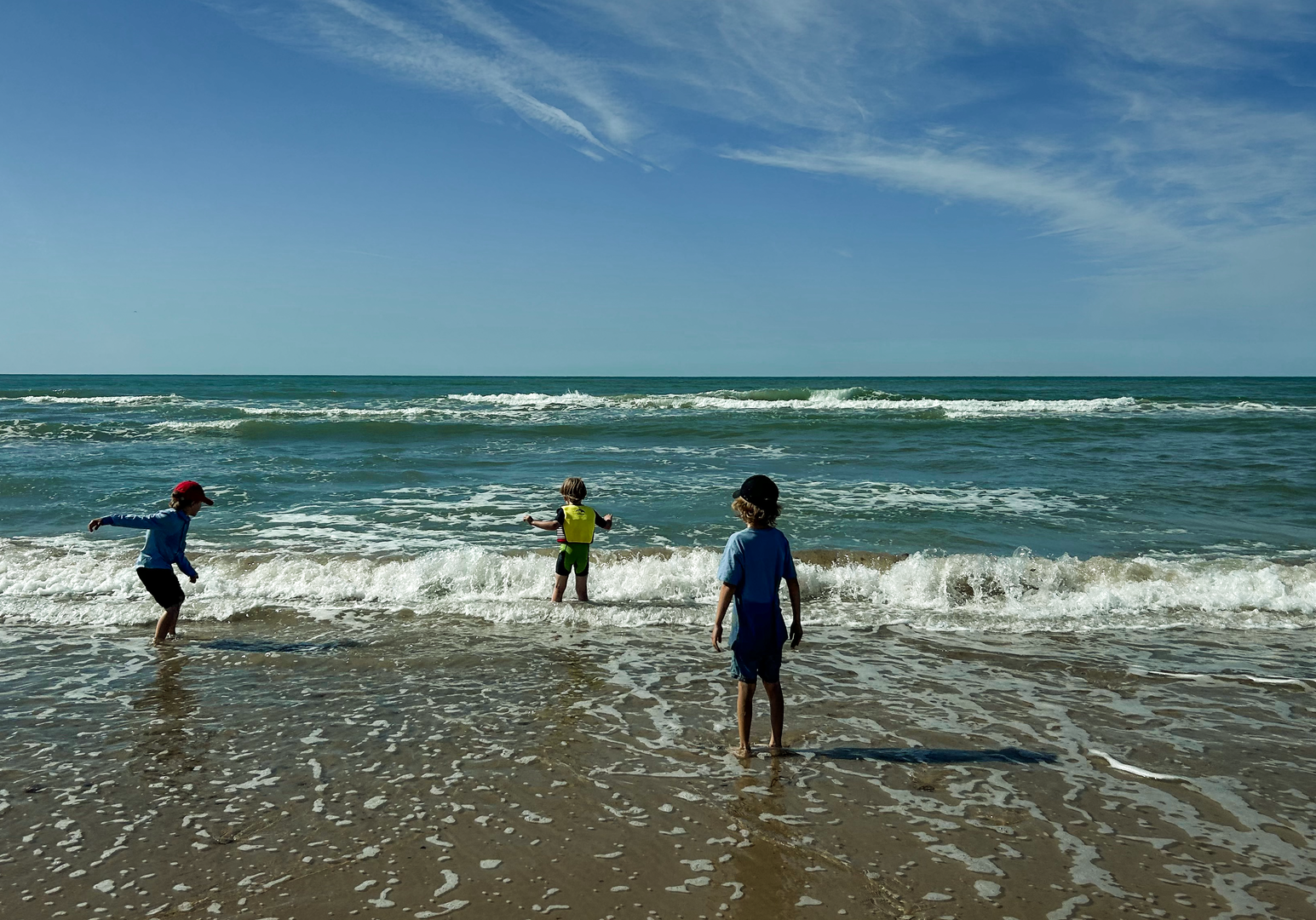 Drei Kinder spielen im flachen Wasser am Strand bei sonnigem Wetter und Wellen