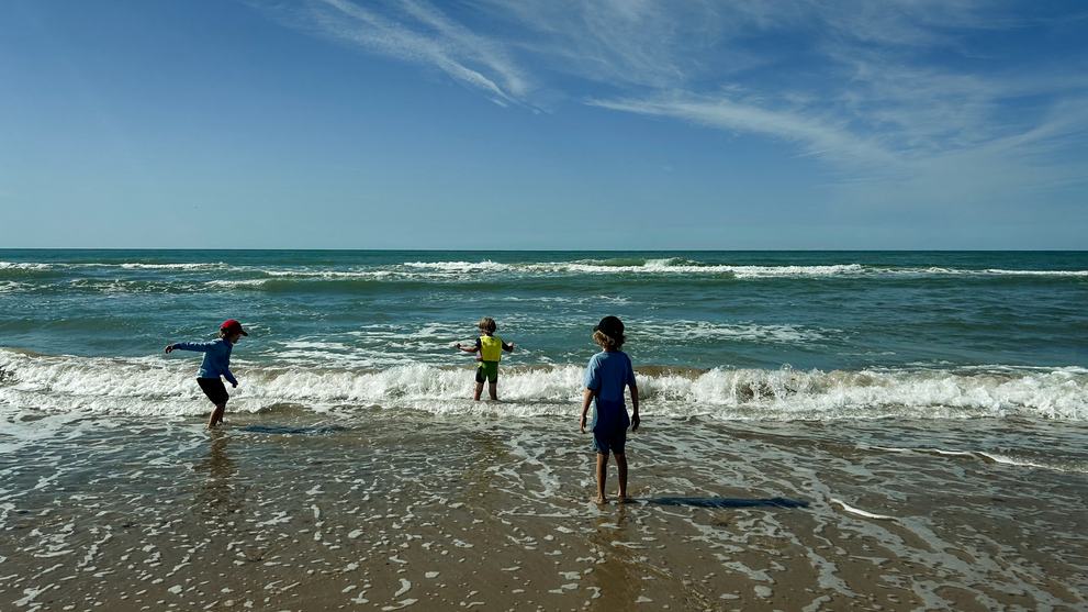 Drei Kinder spielen im flachen Wasser am Strand bei sonnigem Wetter und Wellen