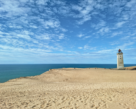 Leuchtturm auf einer sandigen Düne mit Blick auf das Meer unter blauem Himmel