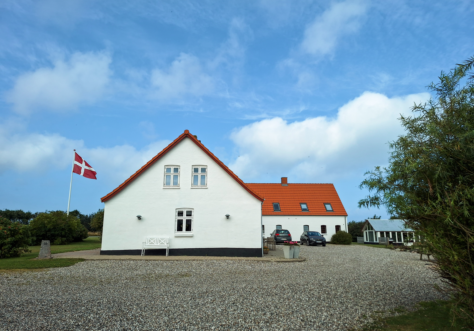 Weißes Haus mit rotem Dach auf einem Hof mit Kiesfläche und Fahnenmast vor blauem Himmel