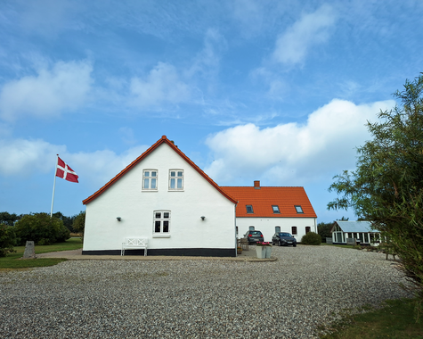 Weißes Haus mit rotem Dach auf einem Hof mit Kiesfläche und Fahnenmast vor blauem Himmel