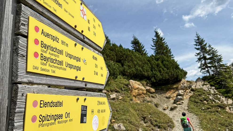 Gelbe Wegweiser an einem Wanderweg in den Bergen mit Personen im Hintergrund