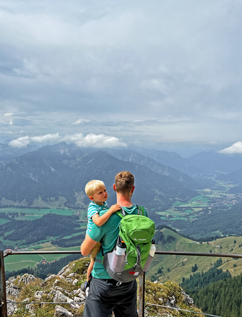 Vater mit Kind auf dem Arm stehen auf einer Aussichtsplattform auf einem Berggipfel und genießen den Ausblick über ein weites Tal