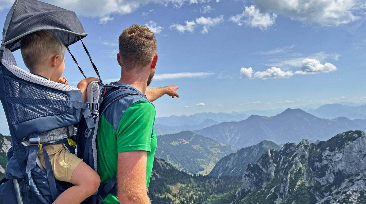 Vater trägt Kind in Kraxe und blickt über eine beeindruckende Berglandschaft
