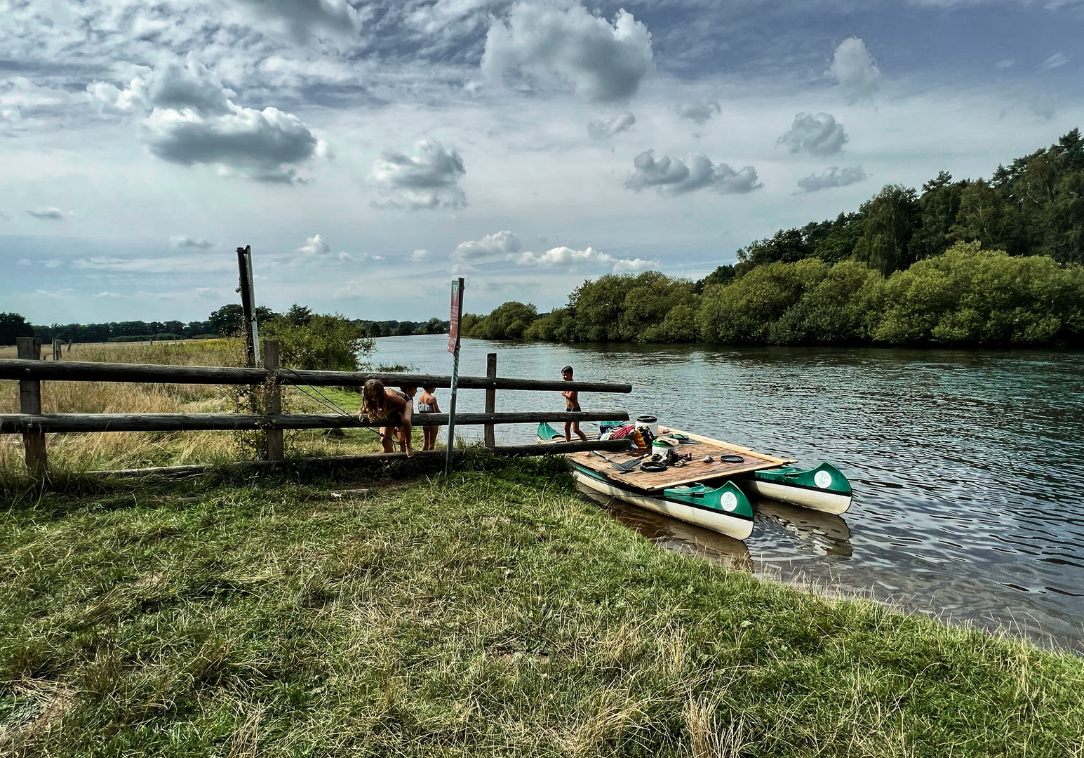 Fluss mit kleinem Steg und zwei Kanus  am Ufer, davor Wiese und Holzzaun und im Hintergrund Bäume und Wolken 