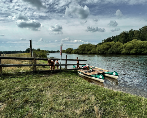 Fluss mit kleinem Steg und zwei Kanus  am Ufer, davor Wiese und Holzzaun und im Hintergrund Bäume und Wolken 