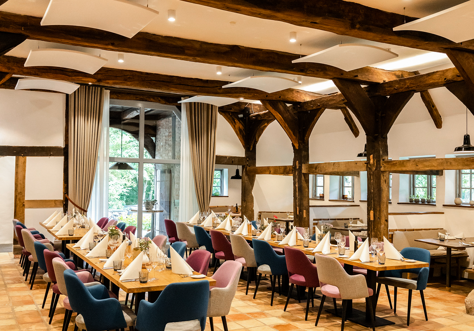 Heller Gastraum mit zentralem Tisch, großen Fenstern und sichtbaren Holzbalken