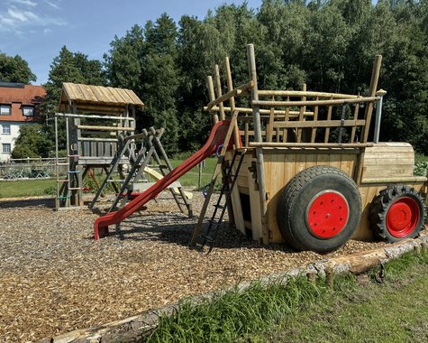 Abenteuerspielplatz aus Holz mit Klettergerüst, Rutsche und großen roten Rädern auf Kiesfläche