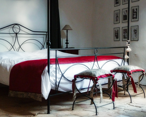 Metallbett mit roter Decke, Hocker und Bilderrahmen an der Wand