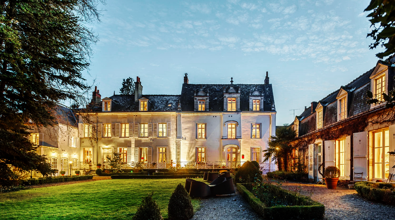 Beleuchtetes Schloss am Abend mit gepflegtem Garten und angrenzenden Gebäuden