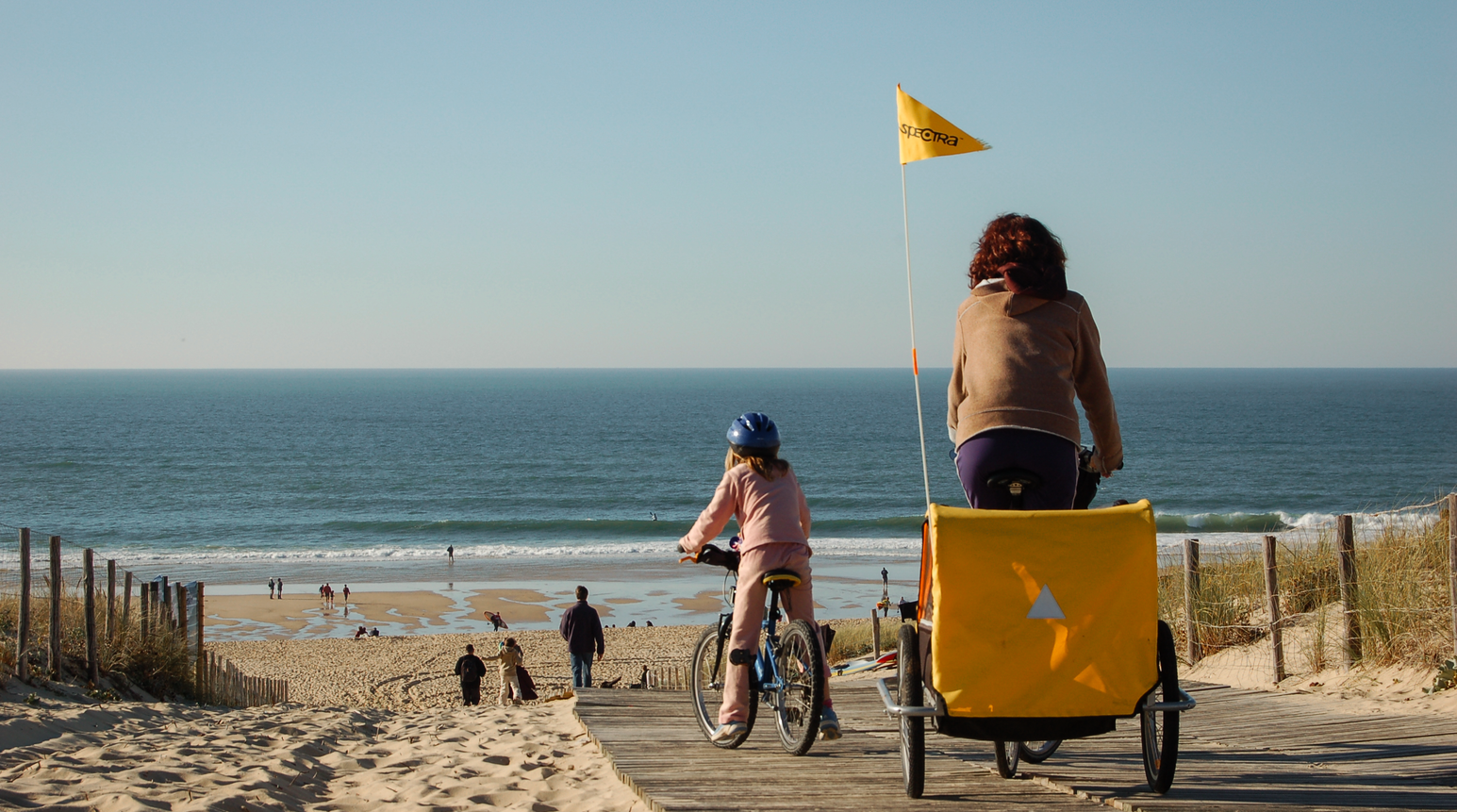 Zwei Personen mit Fahrrad und gelbem Anhänger auf einem Weg durch die Dünen am Meer