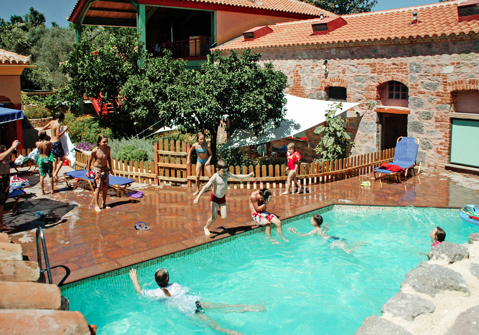 Poolbereich einer Ferienanlage mit Badegästen, Liegen und Gebäuden im Hintergrund.