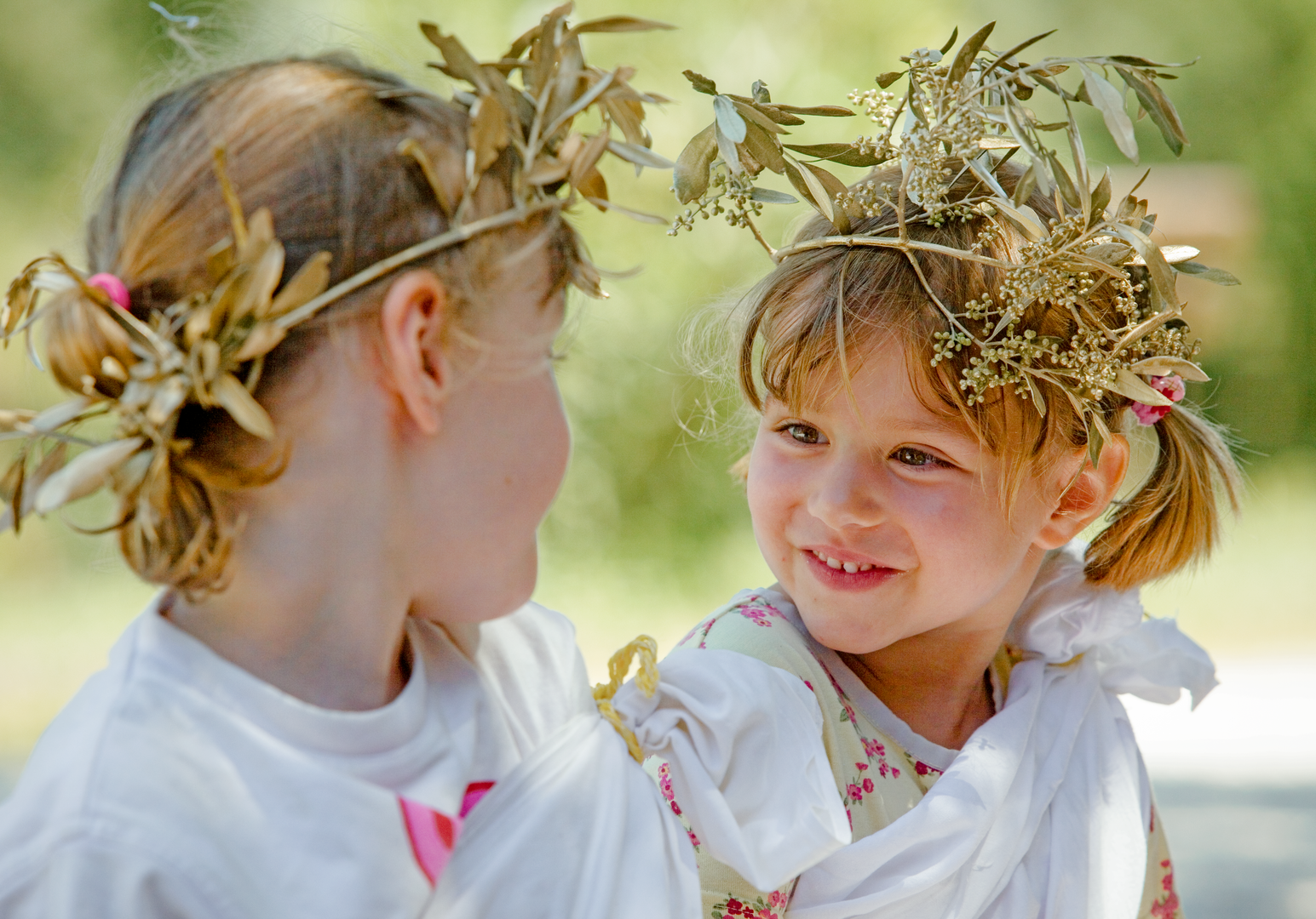 Zwei Kinder tragen selbstgemachte Blumenkränze und lächeln sich im Freien an.
