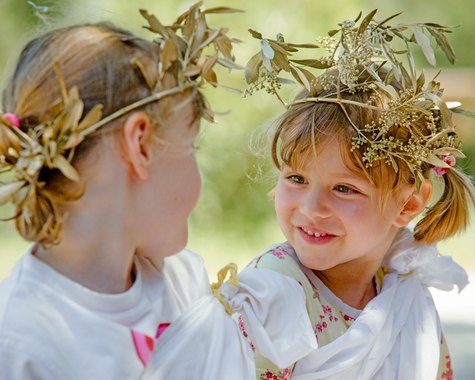 Zwei Kinder tragen selbstgemachte Blumenkränze und lächeln sich im Freien an.