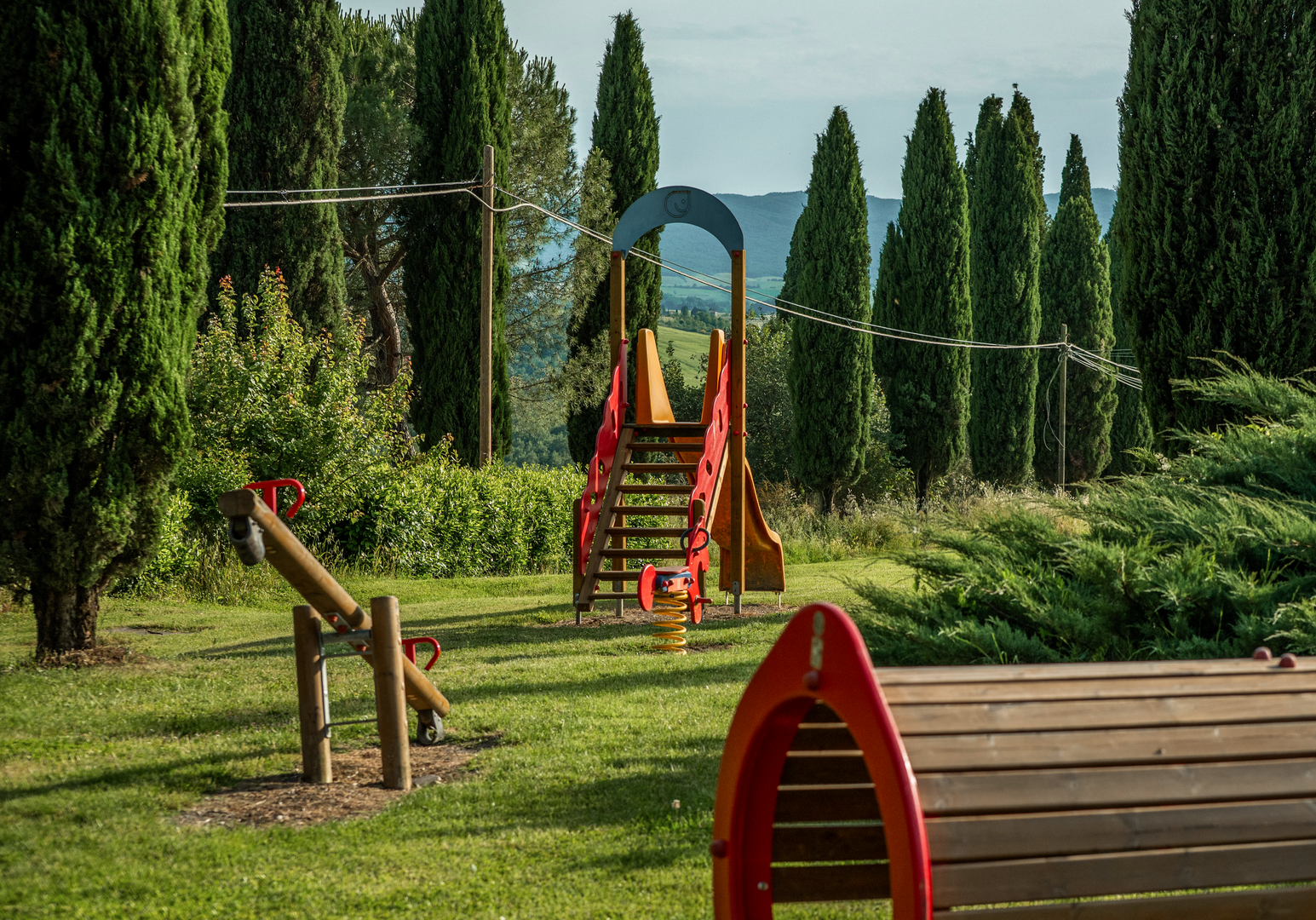 Spielplatz mit Klettergeräten und Rutschen auf einer Wiese, umgeben von Zypressen.
