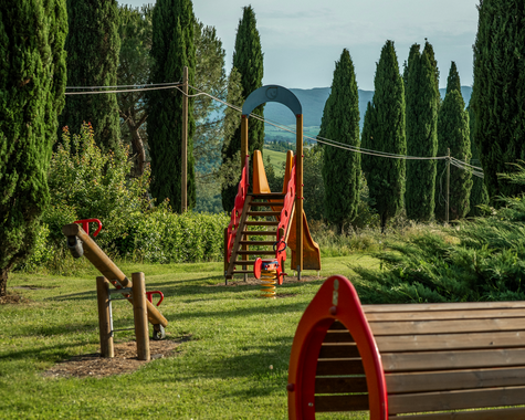 Spielplatz mit Klettergeräten und Rutschen auf einer Wiese, umgeben von Zypressen.