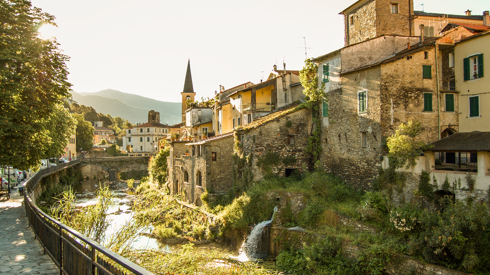 Malerischer Ort mit historischen Häusern entlang eines Flussufers in einer grünen Hügellandschaft.