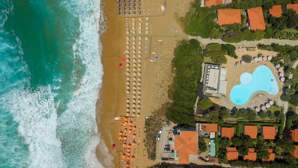 Luftaufnahme einer Ferienanlage mit Pool, Palmen und direktem Zugang zum Sandstrand am Meer.