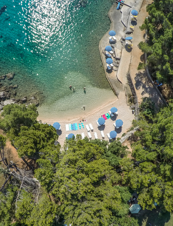 Luftaufnahme einer geschützten Badebucht mit hellem Kiesstrand und klarem Wasser.
