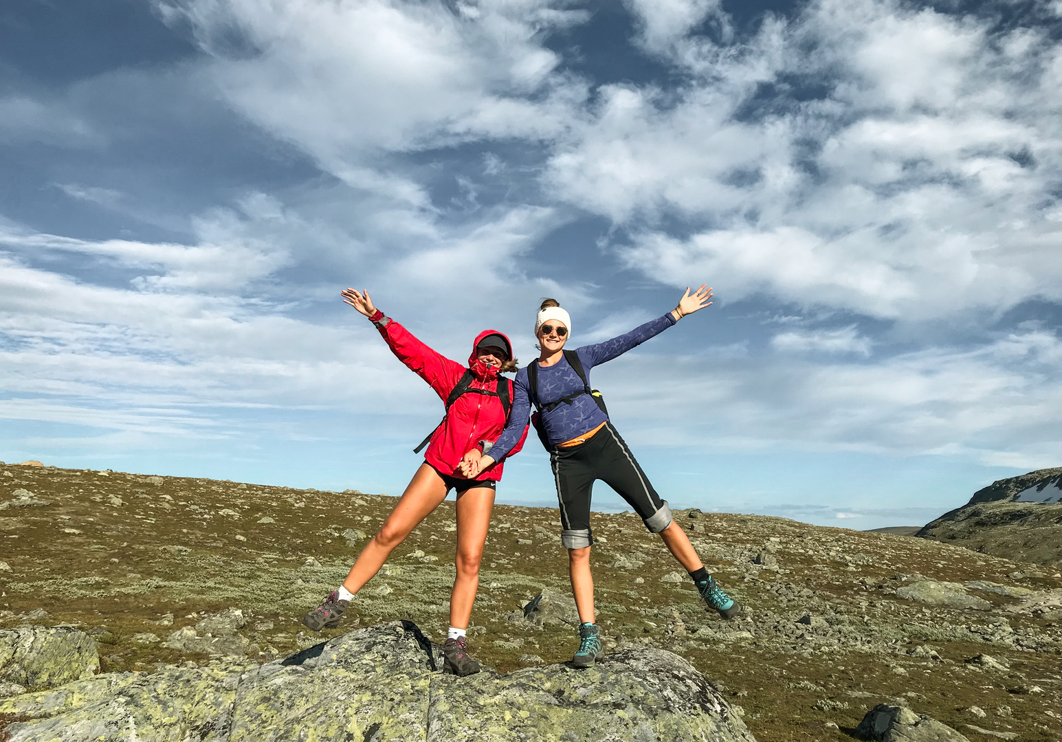 Zwei Wandernde posieren auf einem Bergplateau vor bewölktem Himmel.