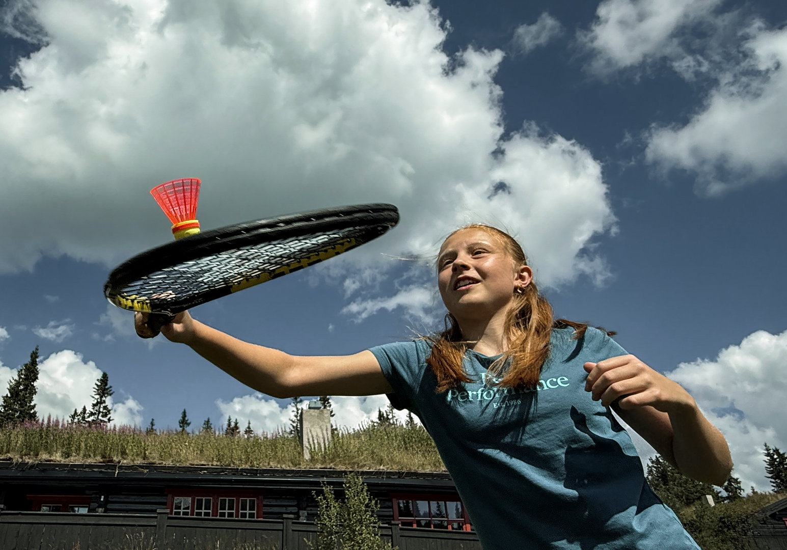 Kind spielt mit einem Tennisschläger auf einer Bergwiese unter wolkigem Himmel.