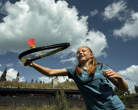 Kind spielt mit einem Tennisschläger auf einer Bergwiese unter wolkigem Himmel.