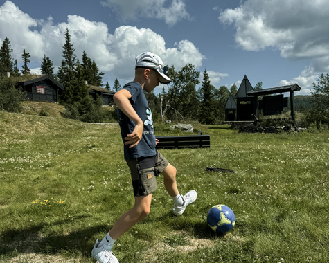 Kind balanciert einen Fußball auf einer Wiese unter wolkigem Himmel.
