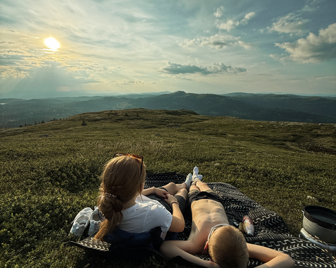 Personen entspannen auf einer Wiese mit Blick auf einen See und die umliegende Berglandschaft.